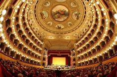 an auditorium filled with lots of people sitting on the stage and looking up at the ceiling