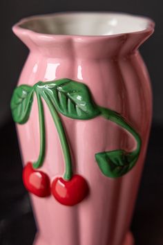 a pink vase with two cherries painted on the front and sides, sitting on a black surface