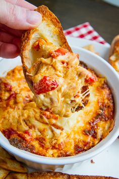 a hand holding a piece of bread over a bowl of cheesy pizza dip