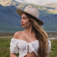 a woman with long hair wearing a white top and hat standing in front of mountains