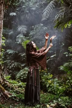 a woman standing in the middle of a forest holding her hands up to the sky