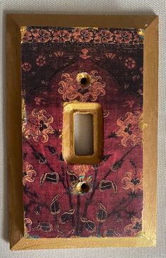 a light switch cover with an ornate design and gold trim on red fabric, framed in wooden frame