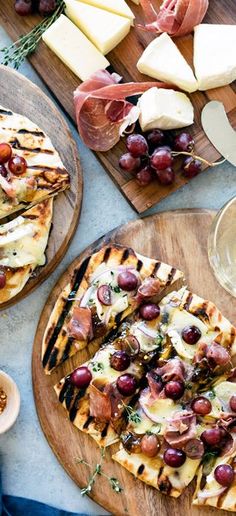 two pizzas on wooden plates with cheese, grapes and other appetizers next to them