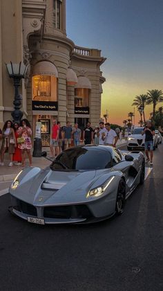 a silver sports car parked in front of a tall building with people standing around it