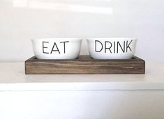 two white bowls with the words eat drink on them sitting on a wooden tray in front of a window