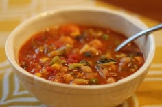 a white bowl filled with vegetable soup on top of a yellow table cloth next to a spoon