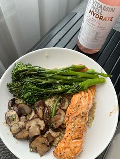 a white plate topped with salmon, mushrooms and broccoli next to a bottle of wine