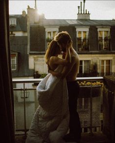 a man and woman standing next to each other on top of a balcony with buildings in the background