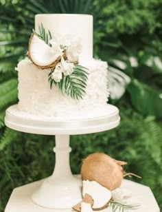 a white wedding cake with coconuts and flowers on the top is surrounded by greenery