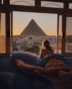 a woman sitting on top of a bed in front of a window next to a pyramid