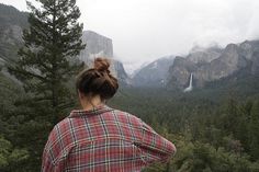 a woman standing on top of a lush green forest next to tall rocks and trees