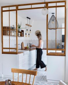 a woman walking through a kitchen next to a table with chairs and shelves on the wall