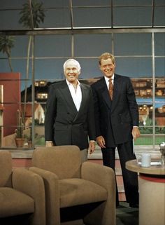 two men standing next to each other in front of a tv set with chairs and couches