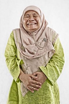 an older woman in a green dress and head scarf smiles while standing against a white wall