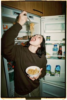 a woman eating cereal from a bowl in front of an open refrigerator