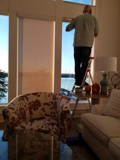 a man standing on a ladder in front of a window next to a couch and chair