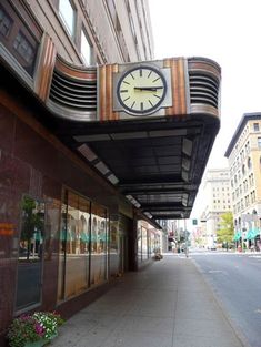 a large clock on the side of a building next to a sidewalk with flowers in front of it