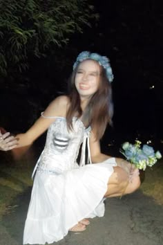 a woman in a white dress kneeling down with flowers on her head and hands out to the side