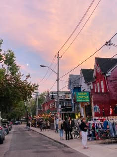 people are standing on the sidewalk in front of shops and parked cars at sunset or dawn