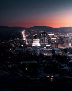 the city is lit up at night with lights shining on buildings and mountains in the background