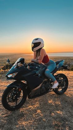 a woman sitting on top of a motorcycle in front of the sun setting over water