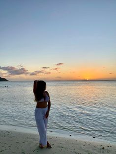 a woman standing on top of a sandy beach next to the ocean at sun set