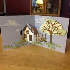 an open card with a house and tree in the middle on a table next to a lamp