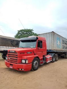 a red semi truck parked in front of a shipping container on the side of a dirt road