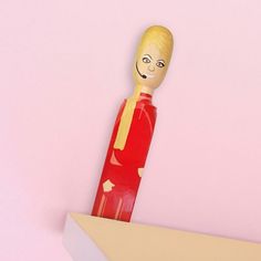 a wooden doll is standing on top of a book shelf with a pink wall in the background