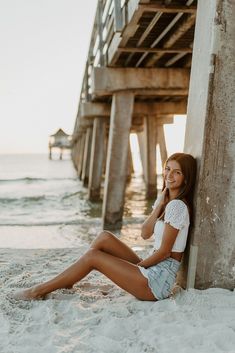 a beautiful young woman sitting on top of a sandy beach next to the ocean under a pier