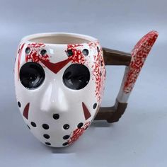 a white and red mask mug with two black holes on it's face, sitting next to a brown handle