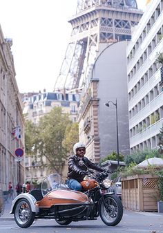 a man riding on the back of an orange motorcycle down a street next to tall buildings