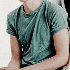 a young man sitting on top of a chair next to a white wall and wearing a green shirt