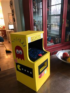 a yellow arcade machine sitting on top of a wooden table next to a bowl of fruit