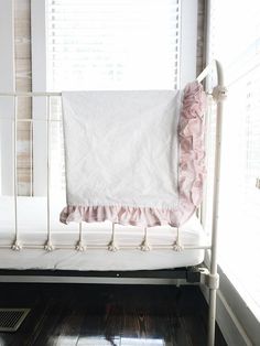 a white crib with a pink ruffled pillow on it's side and a window in the background