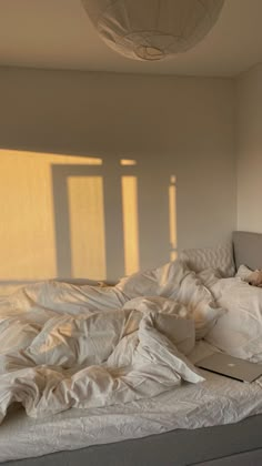 an unmade bed with white sheets and a laptop on it, in front of a window