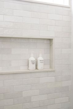 two bottles of shampoo sitting on a shelf in a white tiled bathroom with no shower curtain