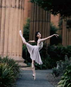 a ballerina is posing in front of some columns and trees with her arms stretched out