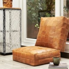 a brown leather chair sitting in front of a window next to a vase and book