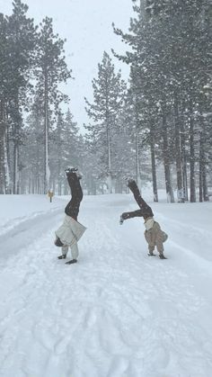 two people doing tricks in the snow near some trees and one person has their arms up