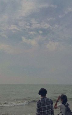 two people standing on the beach looking out at the ocean while one person is holding a camera