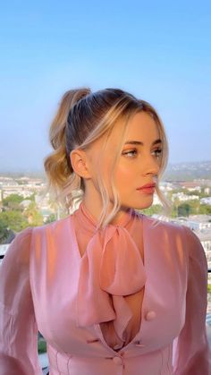 a woman with blonde hair wearing a pink dress and looking off into the distance while standing in front of a cityscape