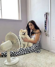 a woman sitting on the floor holding a small white dog next to an electric fan