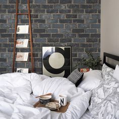 an unmade bed with white sheets and pillows in front of a gray brick wall