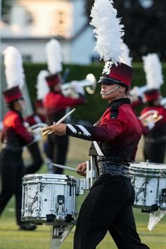 a marching band in red and black uniform with white feathered headdress playing drums