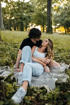 a man and woman sitting on top of a blanket in the grass with trees behind them