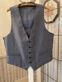 This classic vest looks grey from a distance, but up close it has black, white and medium blue fibres in the polyester blend fabric. It has the original buttons and two front pockets. The back and lining are made from silky bluish grey fabric. The measurements, taken with the vest lying flat, are: shoulder to shoulder, 13 inches; armpit to armpit, 22 inches; length, 25 inches in front and 21 inches in back; bottom edge, 22 inches. In very good condition. Affordable Gray Fitted Vest, Gray Sleeveless Vest Outerwear, Classic Gray Vest For Work, Fitted Gray Vest For Business, Gray Fitted Vest For Business, Classic Gray Formal Vest, Elegant Sleeveless Gray Vest, Classic Tailored Gray Vest, Fitted Gray Vest For Workwear