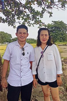 two people standing next to each other in front of a tree and dirt ground with grass