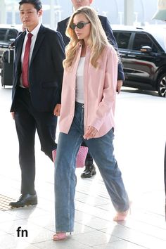 a woman in pink jacket and jeans walking down the street with other people behind her