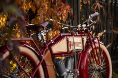 an old fashioned red motorcycle parked in front of a fence with autumn leaves on it